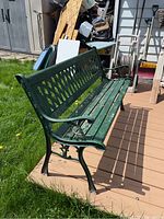 Side view of a green metal frame, wood slatted bench showing diamond pattern backrest and curved armrest with wear on the painted wood slats.