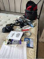 Overview of camera kit on mattress showing camera body with lens attached, charger, cables, manuals, memory card, tripod box, and backpack