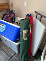 Photo showing two hard coolers (blue and gray), two camping chairs in green and red carrying bags, a purple soft cooler bag, and a lantern near them.