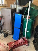 Photo showing the blue padded car creeper leaning against a green storage bin with a red hydraulic floor jack on the floor in front.