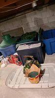 Wide view of basement crawl space showing stacked storage totes, green tree stand lid, artificial tree parts and holiday decorations on white storage bin lid