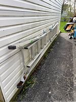 Full-side view of the aluminum extension ladder leaning against a light-colored vinyl siding exterior wall, showing overall length and condition.