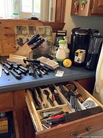 Overall view of the countertop showing the knife block, loose knives, cutting boards and drawer open with kitchen utensils visible. Also includes various cutting tools spread out.