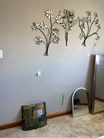 Full view of three floral metal framed wall art pieces mounted on wall and curved metal abstract wall decor panel resting on the floor leaning against the wall.