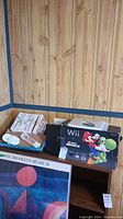 View of original Nintendo Wii box, game cases in box, and controllers on wooden surface against wood panel walls