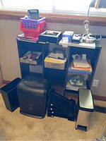 Full view of brown wood shelf with nine cubbyholes filled with various office supplies, black file holders, plastic bins, papers, and a silver desk lamp. Wastebaskets and black office organizers in front of the shelf.