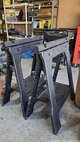 Two black resin Blue Hawk folding sawhorses shown in folded position on garage floor with visible dirt and dust.