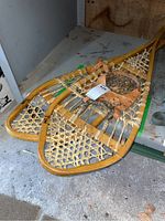 Pair of wooden frame snowshoes with rawhide webbing and leather bindings laid side by side on a shelf, showing full length and condition.