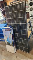 Photo of a large blue-black solar panel with white grid lines, standing upright next to its box, placed on a workshop floor.