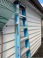 Full view of the blue fiberglass Werner 6 foot step ladder with black top and aluminum steps leaning against a white siding wall.