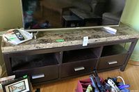 TV bench with faux stone laminate top, three drawers with silver handles, and open shelving beneath. TV sits on top with remote and some items on shelf beneath.