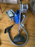 Blue Miele Dimension Plus S2 canister vacuum with hose and metal extension wand connected to floor head, manual and various attachments in plastic bin nearby.