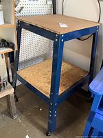 Full view of metal framed work table with plywood top and lower shelf on casters showing blue painted legs and visible wear on wood surfaces.