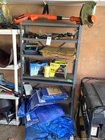 Full view of the metal shelving with Black and Decker electric trimmer on top, gardening tools on shelves, stacks of tarps and bags on floor and shelves.