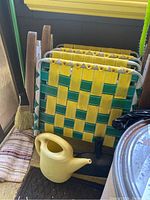 Four folded vintage lawn chairs with yellow and green woven webbing and wooden armrests leaning against a window sill, showing their profiles including the aluminum frames and fastening rivets on the straps.