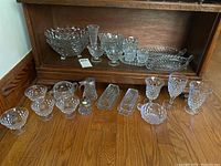 Full collection of Fostoria glassware arranged on wooden floor, showing ice cream bowls, butter dishes, wine glass, vase, syrup jar, sugar dish, candy dishes, cream and sugar dish.