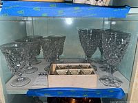 Five water glasses and five wine glasses made of Fostoria crystal with textured geometric pattern, displayed next to a box containing six small crystal salt cellars.
