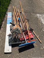 Photo showing a collection of garden tools including rakes, shovels, roof snow rake, hoe, limb trimmer and hole digger laid out on pavement.