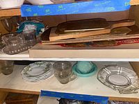 Shelf with clear glass cups, glass plates, cream and sugar set on tray, MCM wooden lap trays, and assorted glassware.