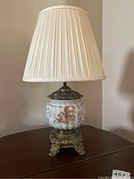 Full view of the lamp showing pleated beige shade, floral glass body, and metal base on wooden surface.