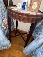 Round wooden side table shown with decorative carved pattern on the edge of the tabletop and an X-shaped stretcher base connecting the legs. Visible scratches on surface.