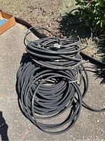Four coiled black soaker hoses shown outdoors on pavement with some greenery and a cardboard box corner visible.