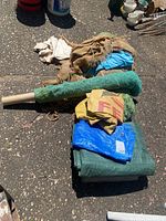 Overall view of all items showing stacked plastic tarps, burlap sacks, and garden fabric rolled up on pavement.