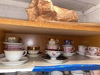 Shelf of vintage porcelain teacups and saucers in various styles and patterns, primarily floral and gold accents.