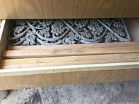 Photo of drawer with stacked carved wooden decorative panels featuring leaf and floral designs, partially visible metal garden decoration.