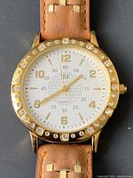 Front of the ladies watch showing the gold tone case with crystal accents and white dial with gold markers, tan leather strap.