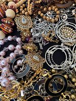 Close-up photo showing assorted vintage jewelry including bead necklaces, gold-tone and silver-tone earrings with rhinestones, chain link bracelets, and black enamel pieces.