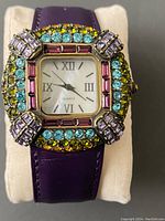 Front view of ladies quartz watch with multicolored decorative stones on bezel and purple leather strap, displayed on a white watch pillow.