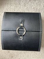 Closed view of compact black faux leather jewelry organizer with metal ring closure and white stitch detail.