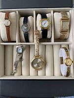 Box with seven vintage ladies watches arranged in a compartmented beige watch storage box, showing a variety of styles and bands.