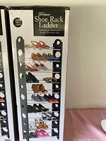 Full view of deluxe shoe rack ladder loaded with shoes against a white wall, showing black frame with circular cutouts on sides and horizontal metal bars holding shoes.