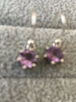 Close-up photo of pair of sterling silver amethyst earrings resting on gray fabric with lever-back closures visible.