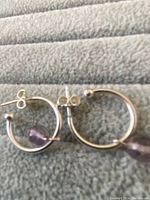 Pair of sterling silver hoop earrings with purple amethyst beads shown lying flat on grey fabric background.