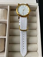 Close-up of the watch face and white woven strap showing gold case and dial with numerals.