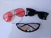 Three pairs of vintage sunglasses displayed on a white background: gold frame aviators with pink lenses, black wayfarers, and black wraparound sunglasses.