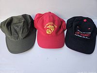Three vintage hats displayed side by side: olive green military-style, red U.S. Marine Corps, and black embroidered 'Surf Dance Off' cap.