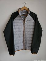 Front view of men's Columbia down filled jacket, light gray body with black sleeves and brown zipper trim, hanging on wooden hanger.