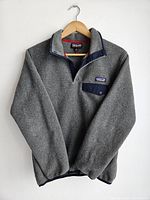 Front view of gray Patagonia Synchilla fleece pullover hanging on wooden hanger, showing snap-button chest pocket and collar with navy trim