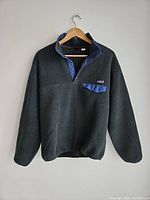 Front view of vintage Patagonia Snap-T fleece pullover hanging on wooden hanger against white wall. Charcoal fleece with blue snap-button placket and pocket flap visible.