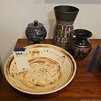 Four African art pottery pieces shown on wooden surface: a large serving bowl, two vases, and a covered bowl with lid.