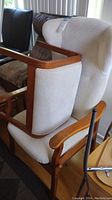 Close-up side angle showing wood armrest, beige patterned upholstery, and back cushion of the chair, with ottoman and other furniture nearby.