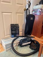 Overview of lot showing Riccar upright vacuum cleaner, Riccar canister vacuum with hose, box of HEPA vacuum bags, and vacuum accessories on floor near door.
