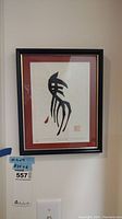 Framed signed and numbered art print hanging on wall, shows black ink abstract design with red mat and black frame.