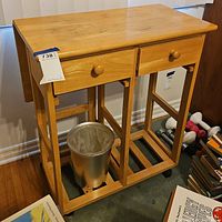 Full view of the wooden storage table showing the two drawers, bottom shelf, and casters with a silver metal trash bin placed on the shelf.
