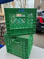 Photo showing two stacked green plastic Neilson milk crates with lattice pattern and handle openings, outdoors near vehicles.
