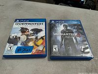 Two PS4 game cases displayed on a flat surface: Overwatch Legendary Edition and Uncharted 4: A Thief's End. A handwritten note labeled '#20 x2' is placed on the Overwatch case.